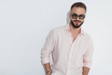 Güneş gözlüğü ve pembe gömlek takan kendine güvenen, elleri ceplerinde beyaz arka planda bekleyen kendine güvenen bir adam.