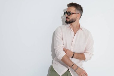 Ciddi bir adam pembe gömlekli ve güneş gözlüklü beyaz arka plana karşı ciddi bir ifadeyle bakarken kollarını sıvıyor.