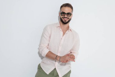 Güneş gözlüklü şık bir adam öne bakıp beyaz arka planda gülümserken ellerini birbirine sürtüyor.