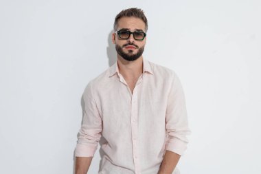 Güneş gözlüğü ve pembe gömlek takan ciddi bir adam beyaz arka planda dikilip ileriye bakıyor.