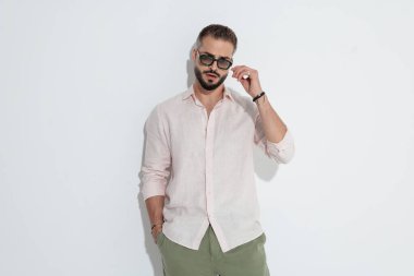 Pembe gömlekli ve yeşil pantolonlu havalı bir adam güneş gözlüğünü ayarlarken el ele durur ve beyaz arka planda ileriye bakar.