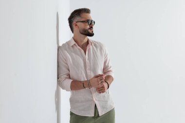 Güneş gözlüğü takan, pembe gömlekli, ellerini birbirine sürterek beyaz arka plana bakan şık bir adamın yan görüntüsü.