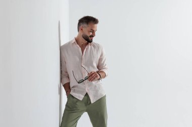 Burun halkalı neşeli adam, duvara yaslanmış gözlük tutuyor ve beyaz arka plana bakarken gülümsüyor.