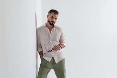 Cebinde güneş gözlüğü olan ve beyaz arka planda ileriye bakarken duvara yaslanan kendine güvenen bir adam.