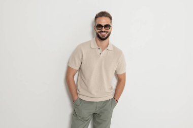 Güneş gözlüğü ve bej örgü polo tişört giyen neşeli sakallı adam elleri ceplerinde gülümseyerek beyaz arka planda tek başına ileriye bakıyor.