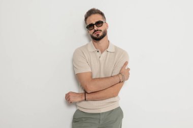 Güneş gözlüğü takan havalı bir adam ve kolları çapraz duran polo tişörtlü beyaz arka planda yalnız başına ileriye bakıyor.