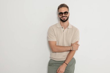 Güneş gözlüğü takan, polo tişörtlü, kolları çapraz gülümseyen ve beyaz arka planda ileriye bakan havalı bir adam.