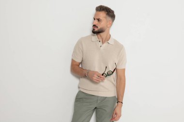 Güneş gözlüğü tutan şık bir adam beyaz arka planda polo tişört giyerken yan tarafa bakıyor.