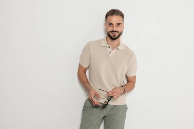 Güneş gözlüğü takan, bej renkli polo tişört giyen ve beyaz arka planda ileriye bakan mutlu bir adam.