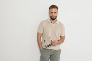 Ciddi bir adam elinde gözlük, cebinde bej polo gömlekle poz veriyor ve beyaz arka planda ileriye bakıyor.