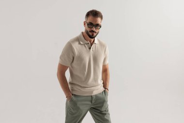 Güneş gözlüğü takmış, elleri ceplerinde duran şık bir adam gri arka planda ileriye bakıyor.