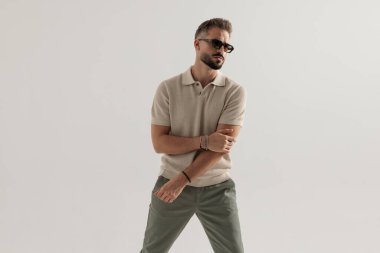 Havalı adam güneş gözlüğü takıyor, bej polo tişört giyiyor, kollarını tutuyor ve gri arka planda izole edilmiş bir yer arıyor.