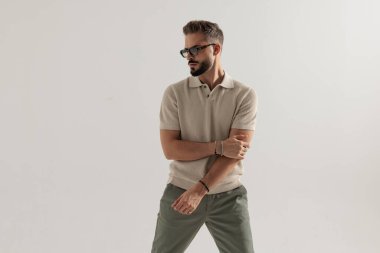 Güneş gözlüğü takan şık bir adam, bej polo tişört giyiyor, kollarını tutuyor ve gri arka planda izole edilmiş bir yer arıyor.
