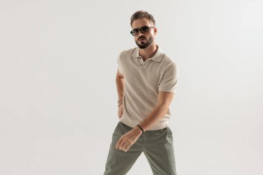 Güneş gözlüğü takan ve bej renkli polo tişört giyen kendine güvenen bir adam öne çıkıyor ve gri arka planda izole edilmiş kameraya bakıyor.