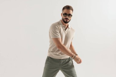 Güneş gözlüğü takan havalı bir adam ve bej polo tişört giyip el ele poz vererek hazırlanıyor ve gri arka planda tek başına ileriye bakıyor.