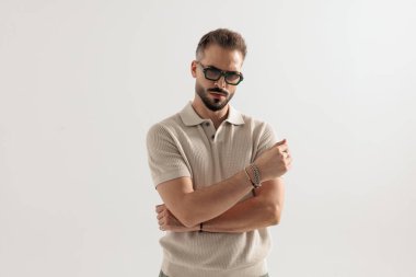 Ciddi bir adam güneş gözlüğü takıyor ve bej polo tişört giyiyor. Kolunu tutuyor ve gri arka planda yalnız başına ileriye bakıyor.