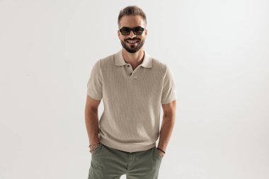Bej renkli polo gömlek ve güneş gözlüğü takan neşeli bir adam elleri ceplerinde ve gri arka planda bekliyor.