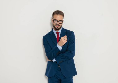 Gözlüklü ve mavi takım elbiseli kendine güvenen işadamı kollarını kavuşturup beyaz arka planda ileriye bakıyor.