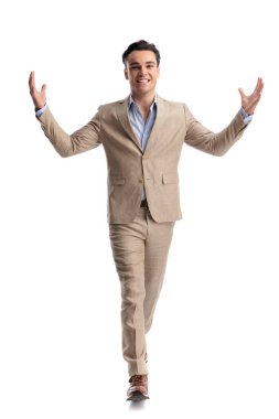 Bej renkli keten bir takım elbise giyen tutkulu bir adam kolları açık yürüyor ve beyaz bir arkaplanda ileriye bakıyor.