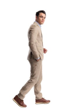 Bej renkli keten bir takım giyen, cebinde yürüyen ve beyaz arka planda izole bir şekilde ileriye bakan şık bir adamın yan görüntüsü.