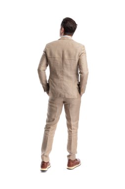 Keten takım giyen, elleri ceplerinde duran ve beyaz arka planda izole bir şekilde başka tarafa bakan zarif bir adamın arka görüntüsü.