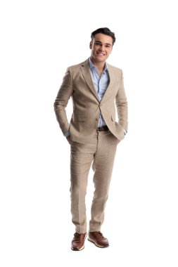Bej renkli keten takım elbiseli mutlu bir adam elleri ceplerinde, beyaz arka planda ileriye bakıyor.