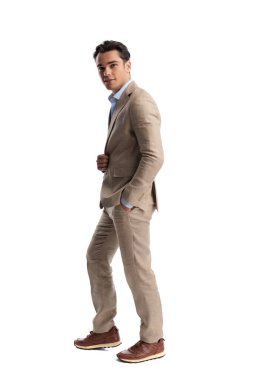 Bej renkli keten takım elbiseli, ceketini cebinde tutan ve beyaz arka planda ileriye bakan kendine güvenen bir adamın yan görüntüsü.