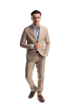 Bej renkli keten takım elbiseli şık bir adam öne çıkıyor ve beyaz arka planda ilerlerken ceketini tutuyor.