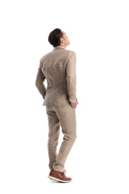 Bej renkli keten takım elbiseli, elleri ceplerinde duran ve beyaz arka plana bakan meraklı bir adamın arka görüntüsü.