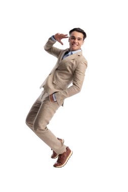 Bej renkli keten bir takım elbise giyen oyuncu bir eli cebinde geriye doğru eğilmiş ve alnına yakın bir el hareketi yaparak beyaz bir arkaplanda ileriye bakıyor.