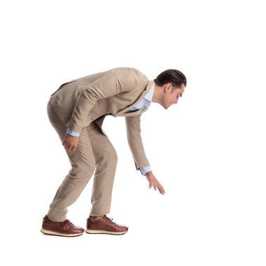 Beyaz bir arka plana bakarken bej renkli keten takım elbiseli meraklı bir adamın öne doğru eğildiğini ve elini aşağıya uzattığını görüyorum.