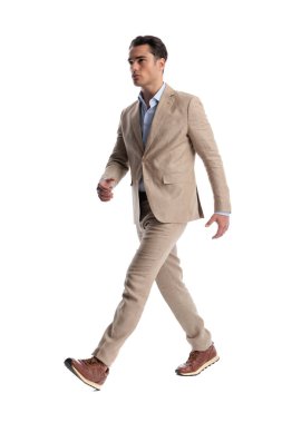 Beyaz arka planda yürüyen bej takım elbiseli ciddi bir adamın yan görüntüsü.