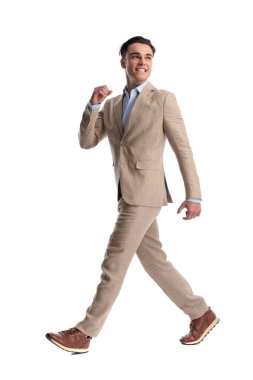 Beyaz arka planda yürüyen ve omzunun üzerinden bakan bej takım elbiseli mutlu bir adamın yan görüntüsü.