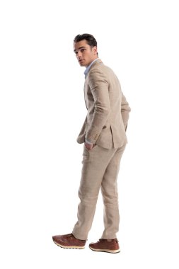 Keten takım elbiseli şık bir adamın arkadan görünüşü elleri ceplerinde yürümesi ve beyaz arka planda izole bir şekilde omzunun üzerinden geriye bakması.