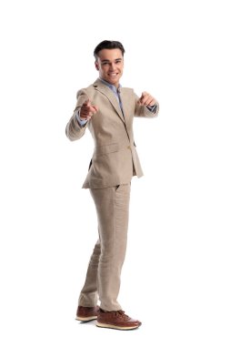 Beyaz arka planda izole edilmiş kameraya bakarken iki işaret parmağıyla ayakta duran ve işaret eden bej keten takım elbiseli mutlu bir adam.