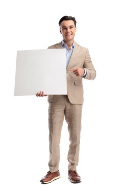 Beyaz, boş bir tahta tutan, gülümseyen ve beyaz arka planda izole bir şekilde ileriye bakan şık keten takım elbiseli mutlu bir adam.