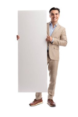 Beyaz arka planda izole bir şekilde ileriye bakarken, beyaz takım elbiseli mutlu bir adam ayakta ve elinde büyük beyaz bir dikey tahta tutuyor.