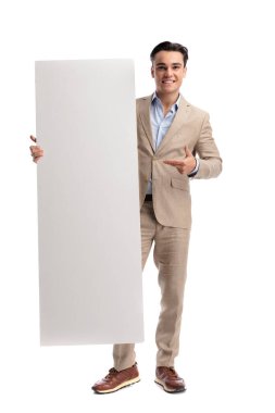 Zeki keten takım elbiseli neşeli bir adam beyaz arka planda tek başına ileriye bakarken dikey boş bir tahtaya bakıp işaret ediyor.