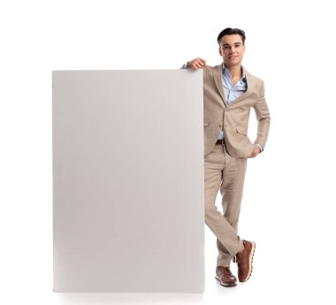 Şık, keten takım elbiseli kendine güvenen, bacak bacak üstüne atmış, büyük, dikey, boş bir tahtaya yaslanmış ve beyaz arka planda tek başına ileriye bakan bir adam.