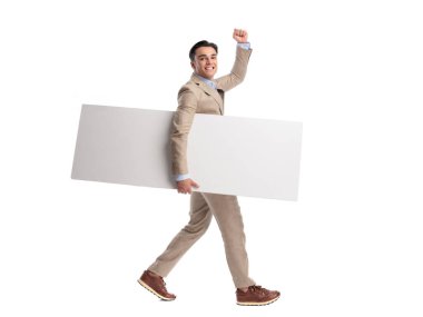 Beyaz arka planda zaferi kutlarken bej renkli keten takım elbise giyen, elinde kocaman boş bir tahtayla yürüyen hevesli bir adamın yan görüntüsü.