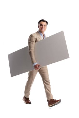 Bej renkli keten takım elbiseli mutlu bir adam beyaz arka planda ilerlerken büyük boş bir tahta tutuyor ve yürüyor.