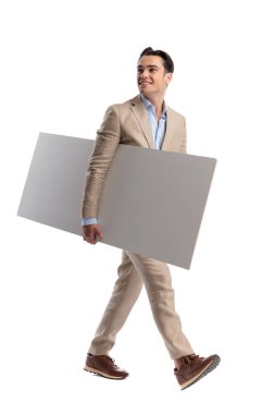 Bej renkli keten takım elbiseli neşeli bir adam beyaz arka plana bakarken elinde kocaman boş bir tahtayla yürüyor.