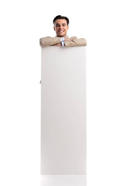 Bej renkli keten takım elbiseli mutlu bir adam beyaz arka plana bakarken dikey boş bir tahtaya yaslanıyor.