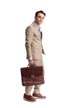 Bej renkli keten takım elbiseli, kahverengi deri çantalı ve beyaz arka planda tek başına ileriye bakan kendine güvenen bir adamın yan görüntüsü.