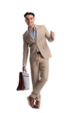 Kahverengi deri evrak çantası tutan ve bacak bacak üstüne atarak beyaz arka planda duran bej renkli keten elbise giyen mutlu bir adam.