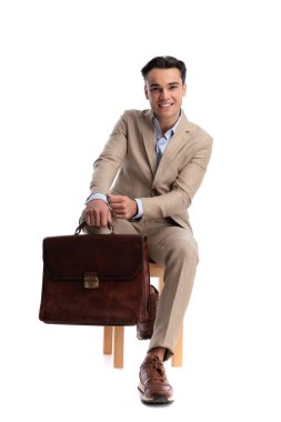 Beyaz arka planda izole edilmiş ahşap sandalyeye otururken elinde kahverengi deri çanta ve kollarını ayarlayan bej renkli keten elbise giyen başarılı bir adam.