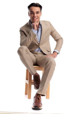 Bej renkli keten bir takım elbise giyen ve çenesine dokunup beyaz arka planda ileriye bakan mutlu bir adam.