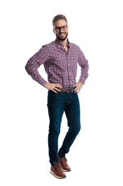 Gözlüklü ve ekose gömlekli neşeli bir adam elleri kalçasında duruyor ve beyaz arka planda izole bir şekilde gülümsüyor.