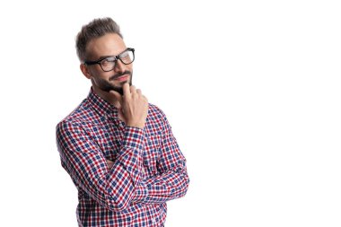 Düşünceli, sıradan, gözlüklü ve ekose gömlekli genç bir adam. Düşünürken ve bakarken çenesine dokunuyor. Beyaz arka planda izole edilmiş.