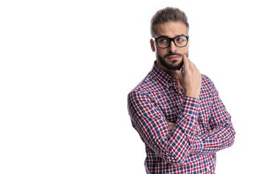 Gözlüklü, kareli tişörtlü, düşüncelere dalmış, beyaz arkaplana bakarken çenesine dokunan düşünceli genç bir adam.